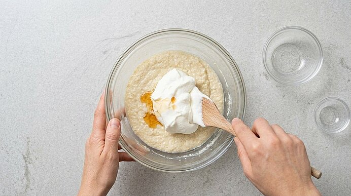 Zwei Hände heben geschlagenes Obers und Orangenlikör mit einem Holzlöffel in einer Glasschüssel unter.