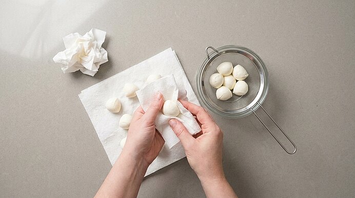 Zwei Hände tupfen Mini-Mozzarellakugeln auf Küchenpapier trocken. Daneben steht ein Sieb mit Mozzarellakugeln.