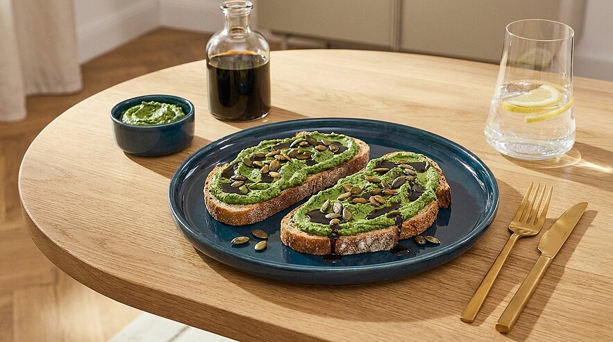 Zwei Scheiben rustikales Brot mit grünem Aufstrich, bestreut mit Kürbiskernen, auf einem blauen Teller.