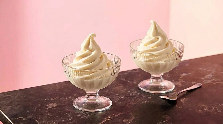 Zwei Glas-Schälchen mit spiralförmig aufgedrehtem Vanille-Softeis auf dunkler Marmoroberfläche, daneben ein Dessertlöffel vor rosa Hintergrund.