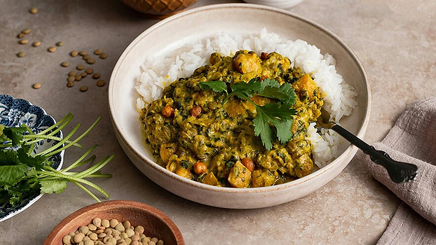 Eine Schüssel mit Reis, in der eine grüne Currysoße mit Gemüse und Linsen serviert wird. Frischer Koriander dekoriert das Gericht. Daneben sind einige grüne Kräuter und eine Schale mit Linsen platziert.