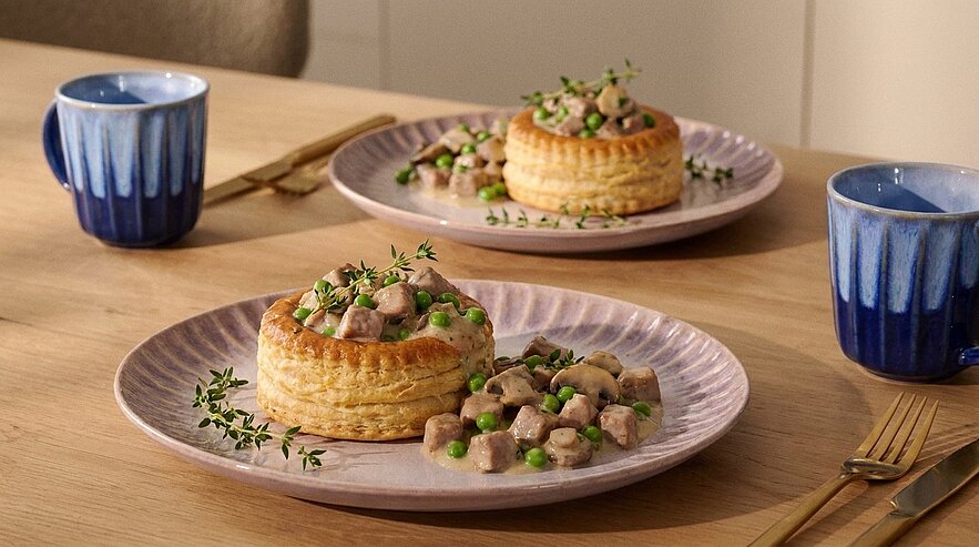Zwei Teller mit Blätterteig-Vol-au-Vent, gefüllt mit cremigem Ragout aus gewürfeltem Fleisch, Champignons und Erbsen, garniert mit frischen Thymianzweigen.