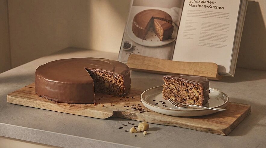 Ein Schokoladen-Marzipan-Kuchen liegt auf einem Holzbrett, mit einem Stück auf einem Teller daneben. Der Kuchen ist mit einer glatten Schokoladenglasur überzogen. Im Hintergrund ist ein Rezeptbuch sichtbar, das den Kuchen zeigt. Schokoladenstückchen sind verstreut.