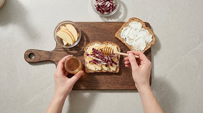 Zwei Hände beträufeln eine mit Ziegenkäse, Apfelspalten und Radicchio belegte Brotscheibe mit Honig.