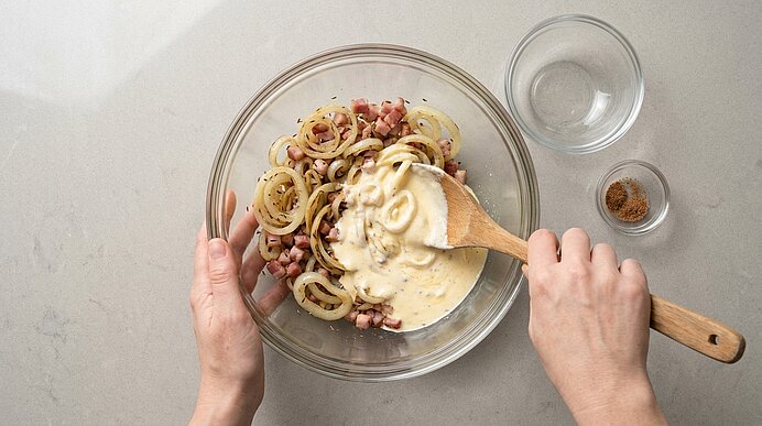 Zwei Hände vermengen mit einem Holzlöffel eine Schmandcreme unter Zwiebeln und Speck in einer Glasschüssel.
