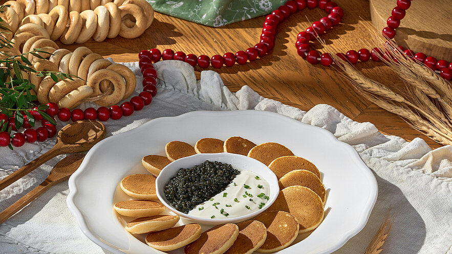 Eine weiße Schale auf einem Tisch, gefüllt mit einem Dip aus saurer Creme und schwarzem Kaviar in der Mitte. Rundherum sind dünne, goldbraune Pfannkuchen angeordnet. Im Hintergrund sind dekorative Elemente wie Gebäck und eine rote Perlenkette zu sehen.