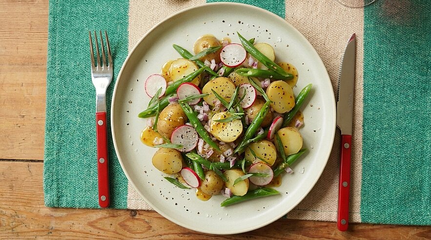 Teller mit Frühlings-Kartoffelsalat aus halbierten neuen Kartoffeln, grünen Bohnen, Radieschenscheiben, fein gehackten roten Zwiebeln, Kräutern und schwarzem Pfeffer.