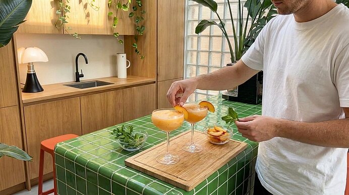 Person garniert zwei orangefarbene Cocktails auf einer Kücheninsel und legt Orangenscheiben als Garnitur auf die Gläser.