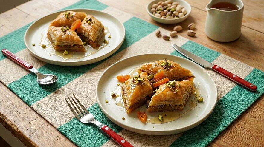 Zwei Teller mit jeweils drei Stücken Baklava, garniert mit gehackten Pistazien und getrockneten Aprikosen, leicht mit Sirup beträufelt.