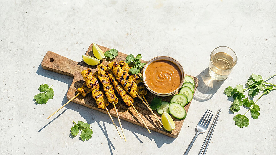 Ein Holzbrett mit gegrillten Fleischspießen, serviert mit Erdnusssauce, Gurkenscheiben und Limettenspalten. Dazu frische Korianderblätter und ein Glas Wasser. Das Gericht vermittelt eine entspannte, sommerliche Atmosphäre.