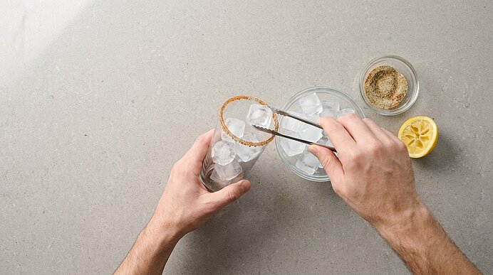 Hände legen mit einer Zange Eiswürfel in ein Glas mit gesalzenem Rand; Schale mit Gewürz und eine halbierte Zitrone liegen daneben.