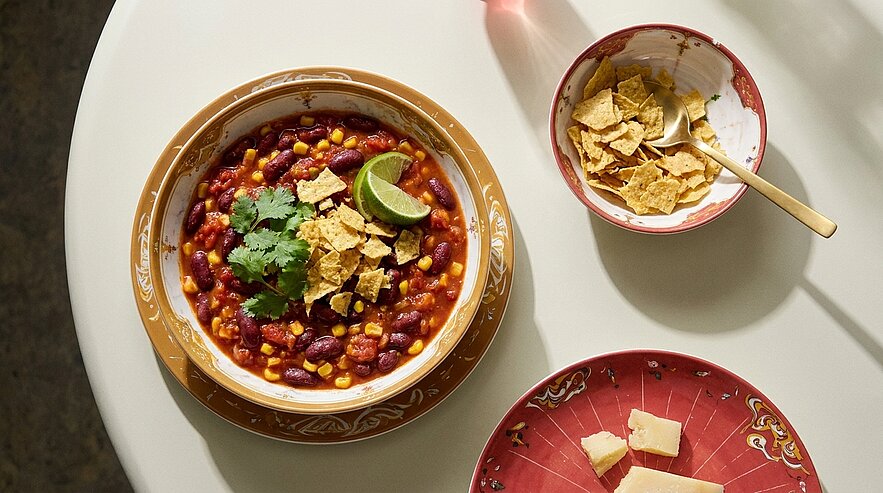 Schüssel mit Bohnen‑und Mais‑Chili in Tomatensauce, garniert mit Koriander, Limettenschnitz und zerbröselten Tortilla‑Chips.
