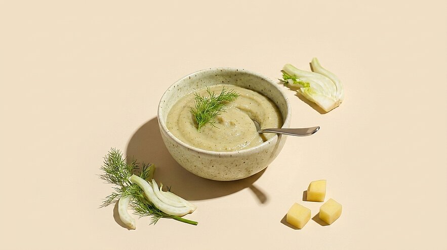 Schale mit grünlicher, cremiger Fenchelsuppe, mit Dill garniert und Löffel darin; daneben Fenchelstücke und kleine Käsewürfel.