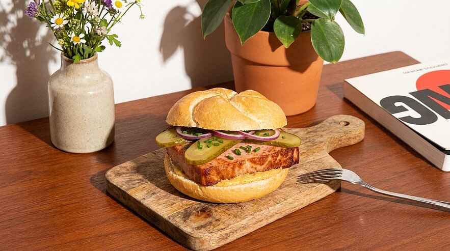 Brötchen mit dickem, gebratenem Fleischstück, Gewürzgurkenscheiben, roten Zwiebelringen und Schnittlauch, serviert auf einem Holzbrett.