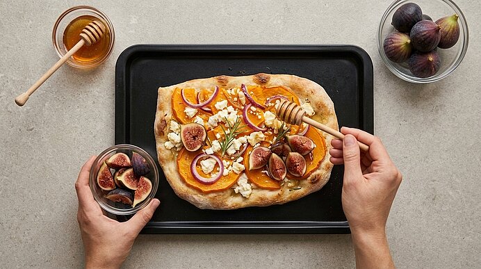 Zwei Hände beträufeln eine Pinsa mit Kürbis, Feta und Feigen mit Honig aus einem Honiglöffel.