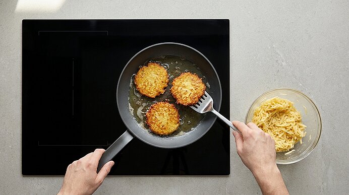 Draufsicht auf zwei goldbraune Kartoffelpuffer, die in einer Pfanne auf dem Herd gebraten werden; eine Hand hält einen Pfannenwender, daneben eine Schüssel mit geriebenen Kartoffeln.