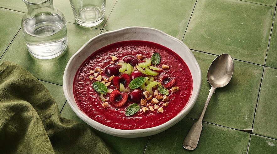 Schale mit tiefroter Kirschsuppe, garniert mit halbierten Kirschen, gehackten Nüssen, Minzblättern und dünnen grünen Scheiben.