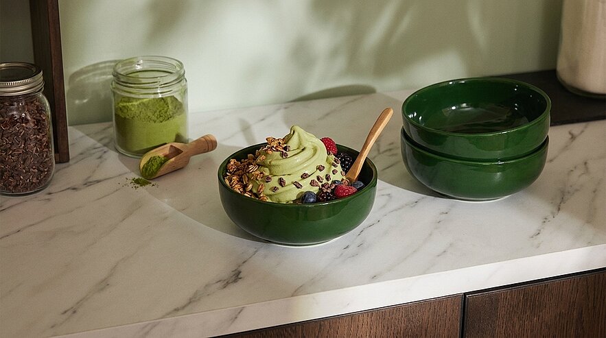 Grün gedrehene Matcha-Softserve in grünem Schälchen, garniert mit Granola, Schokostückchen und frischen Beeren; ein Holzlöffel steckt im Eis.