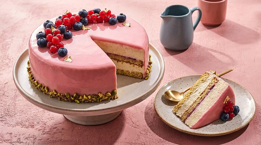 Runde Torte mit rosa Spiegelglasur, eine Scheibe herausgeschnitten. Mehrere Biskuit-Schichten mit Beerenfüllung, dekoriert mit Blaubeeren, roten Johannisbeeren und gehackten Pistazien; ein Stück auf einem Teller daneben.