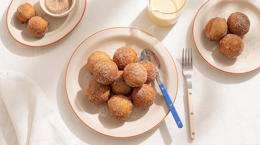 Mehrere zuckerbestreute Quarkbällchen auf Tellern, dazu kleine Schälchen mit Zimt‑Zucker und Vanillesoße; Gabel und Löffel daneben.