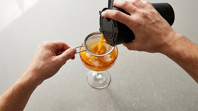 Zwei Hände sieben und gießen einen orangefarbenen Cocktail durch ein feines Sieb in ein Coupéglas mit gewürztem Rand.