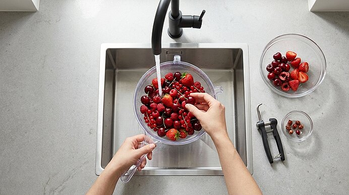 Hände spülen Kirschen und Erdbeeren in einem Sieb unter laufendem Wasser in der Küchenspüle; neben der Spüle Schüsseln mit Beeren und ein Kirschentsteiner.