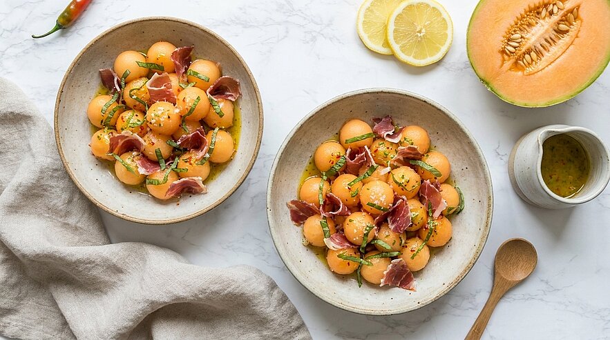 Zwei Schalen mit Melonenkugeln, Prosciutto, frischen Kräutern und Zitronenzesten, dazu ein kleines Gefäß mit Dressing.
