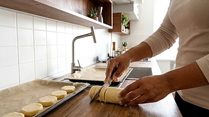 Eine Person schneidet eine Teigrolle in Scheiben; daneben liegen runde Teigstücke auf einem Backblech in einer Küche.