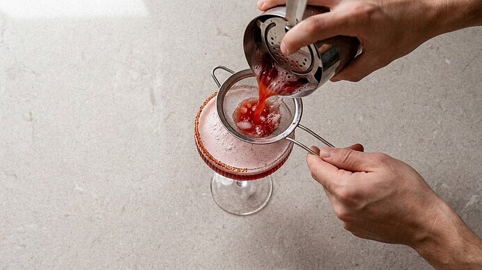 Hände gießen einen roten Cocktail aus einem Shaker durch ein feines Sieb in ein Coupé-Glas.