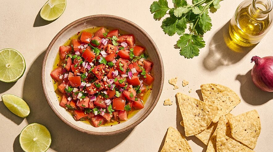 Schale mit frischer Tomatensalsa (gehackte Tomaten, rote Zwiebel, Koriander und Chili), daneben Tortilla‑Chips und Limettenviertel.