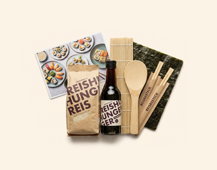 Sushi-Set: Packung Reishunger-Reis, Flasche Reishunger-Essig, Noriblätter, Bambus-Sushi-Matte, Holzlöffel und Essstäbchen im Papierumschlag sowie eine bebilderte Rezeptkarte mit Sushi.