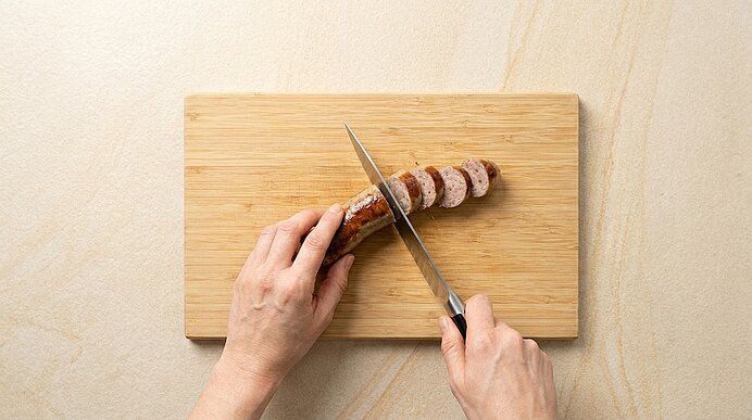Zwei Hände schneiden mit einem Messer auf einem Holzschneidebrett eine Wurst in dünne Scheiben.