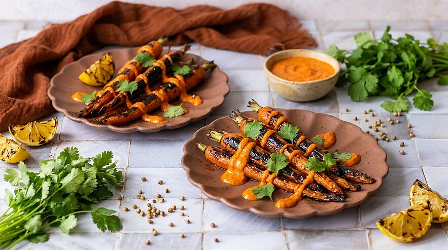 Auf dem Bild sind zwei Teller mit gegrillten Karotten, die mit einer würzigen Sauce und frischem Koriander garniert sind. Daneben befinden sich einige Zitronenscheiben und frischer Koriander. Im Hintergrund ist eine kleine Schale mit Sauce zu sehen.