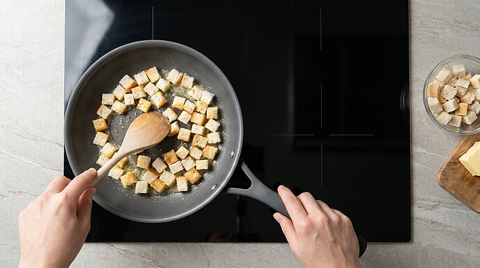Hände rühren mit einem Holzlöffel goldbraun gebratene Brotwürfel (Croutons) in einer Pfanne auf dem Kochfeld.