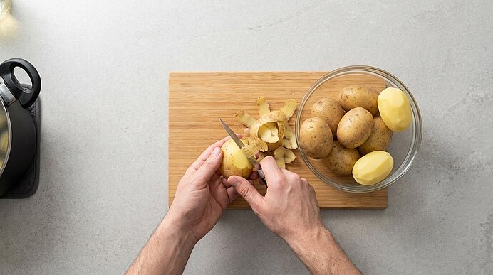 Zwei Hände schälen eine Kartoffel mit einem Messer auf einem Holzbrett; daneben eine Schüssel mit ganzen und geschälten Kartoffeln sowie Kartoffelschalen.