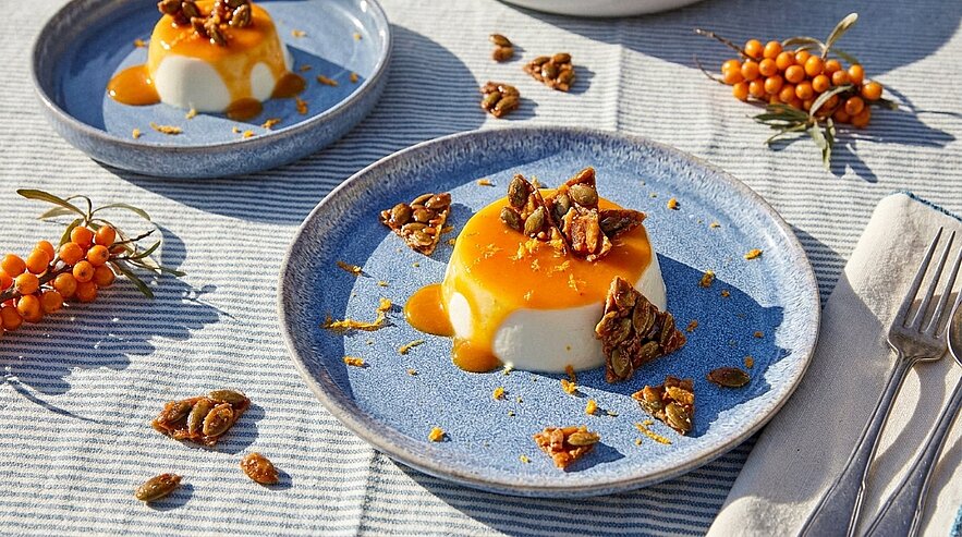 Zwei Teller mit cremigem Panna Cotta, übergossen mit leuchtender oranger Fruchtsoße und garniert mit karamellisierten Nüssen; daneben liegen einige orangefarbene Beerenzweige.