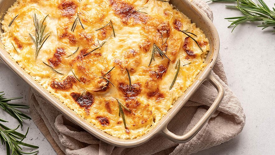 Das Bild zeigt einen köstlichen Kartoffel-Käse-Auflauf, der mit Rosmarin bestreut ist. Der Auflauf ist schön goldbraun gebacken und dampft verführerisch. Neben der Auflaufform liegen frische Rosmarinzweige, die dem Gericht zusätzliches Aroma verleihen. Unter der Auflaufform liegt ein braunes Tuch, das dem Bild eine rustikale und gemütliche Note verleiht.