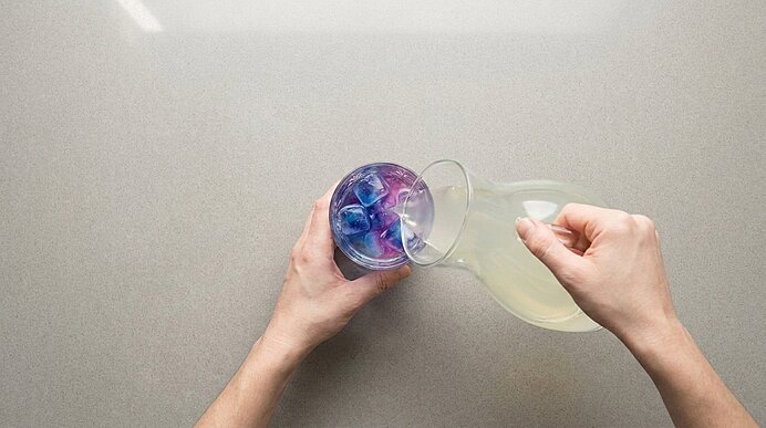 Zwei Hände gießen aus einer Glaskaraffe eine hellgelbe Flüssigkeit in ein Glas, das mit blau‑violetten Eiswürfeln gefüllt ist.