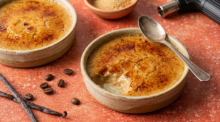Zwei Keramikförmchen mit Crème brûlée und knusprig karamellisierter Zuckerkruste; eines wurde mit einem Löffel angerissen, sichtbar cremige Vanillefüllung.