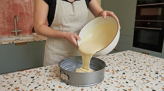 Person in Schürze gießt hellen Kuchenteig aus einer Schüssel in eine runde Springform auf der Arbeitsfläche.