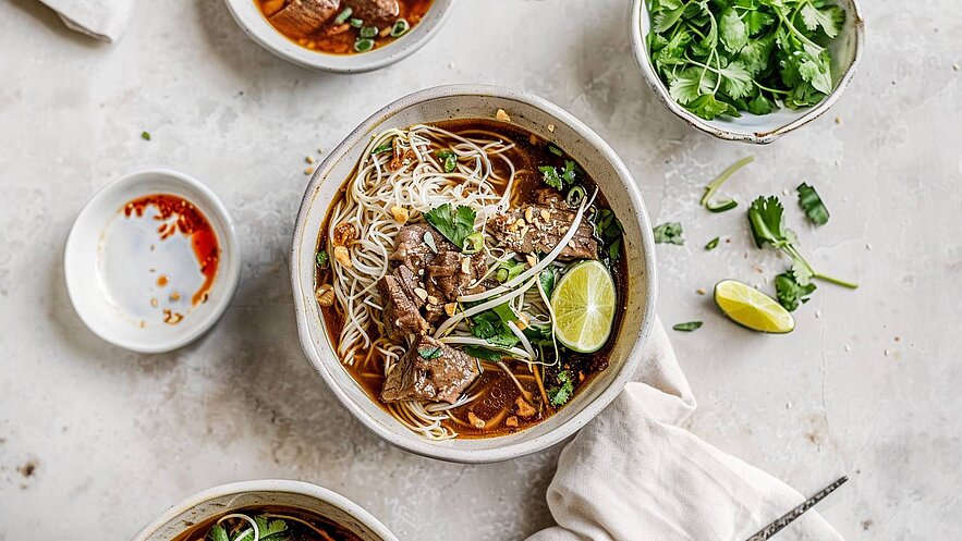 Eine Schüssel mit Nudelsuppe ist im Mittelpunkt, gefüllt mit Rindfleisch, Brühe, frischen Kräutern und Limettenscheiben. Neben der Hauptschüssel stehen kleine Schalen mit Soße und frischem Koriander. Der Untergrund ist hell und schlicht.