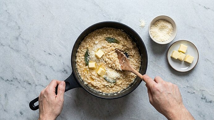 Zwei Hände rühren Butterwürfel und geriebenen Parmesan mit einem Holzlöffel in das Risotto im schwarzen Topf.