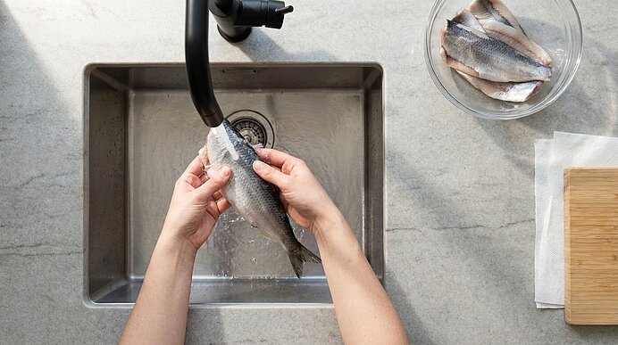 Zwei Hände halten einen ganzen Fisch unter fließendem Wasser über einem Spülbecken; rechts eine Schüssel mit Filets und ein Schneidebrett.