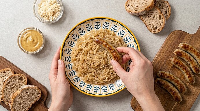 Nahaufnahme: Zwei Hände halten eine gebratene Bratwurst über einem Teller mit Sauerkraut; dazu Senf, geriebener Meerrettich, Brotscheiben und mehrere gebratene Würstchen auf einem Holzbrett.