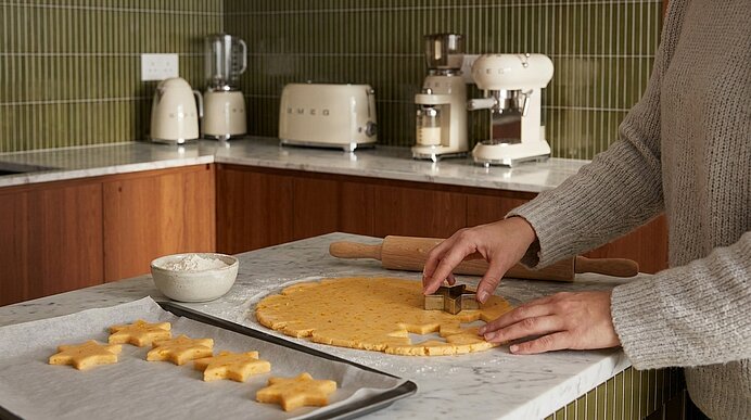 Hände schneiden Sternplätzchen aus ausgerolltem Teig auf einer Marmorarbeitsfläche; daneben Nudelholz, Schüssel mit Mehl und ein Backblech mit ausgestochenen Sternen.