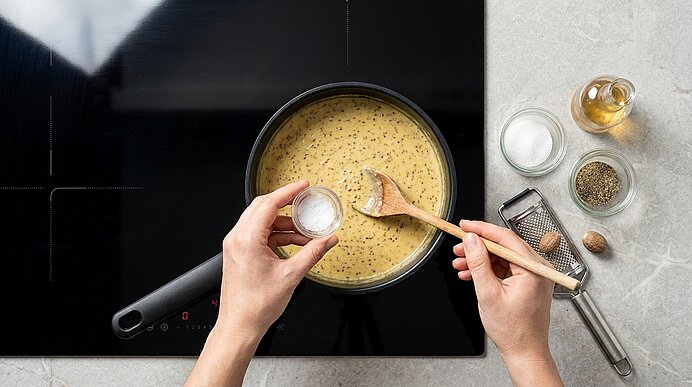 Nahaufnahme: Zwei Hände würzen und rühren mit einem Holzlöffel eine cremige Sauce in einem Topf auf dem Herd; Gewürze und eine Muskatreibe liegen daneben.