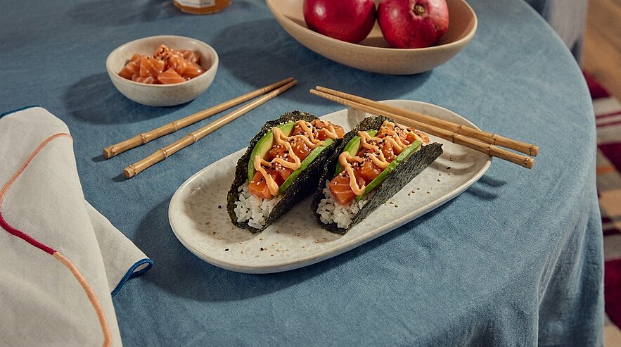 Zwei Sushi-Tacos: Noriblätter gefüllt mit Reis, rohem Lachs, Salatstreifen und würziger Sauce auf einem ovalen Teller.