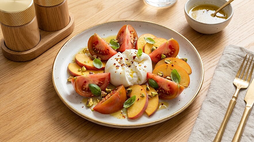 Teller mit Burrata in der Mitte, umgeben von Tomatenspalten und Pfirsichscheiben, garniert mit Basilikumblättern, gehackten Pistazien, Olivenöl und Gewürzen.