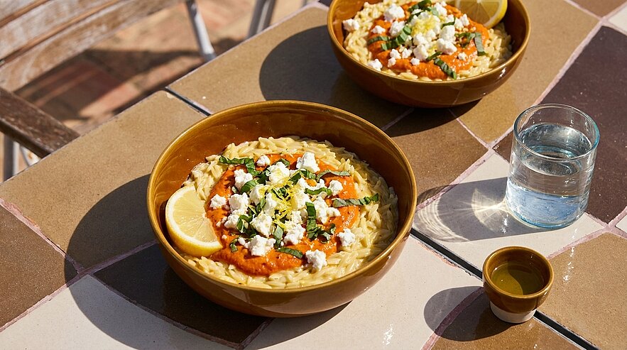 Zwei Schalen mit cremigem Reisgericht, garniert mit Tomatensauce, zerbröseltem Feta, Zitronenscheibe und Kräutern auf einem gefliesten Tisch.