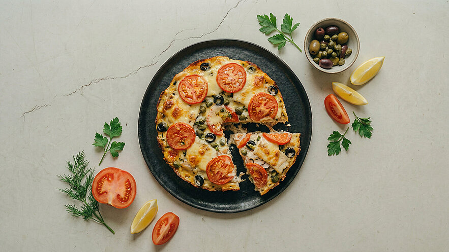 Ein runder, belegter Pizza auf einem schwarzen Teller. Die Pizza ist mit Tomatenscheiben, Oliven und Käse belegt. Ein Stück wurde herausgenommen. Um den Teller herum liegen frische Kräuter, eine halbe Zitrone und einige Kirschtomaten.